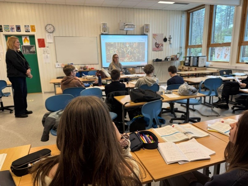 Obraz przedstawia pomieszczenie szkolne, dzieci przy ławkach z książkami, nauczyciel