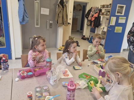 Obraz sali przedszkolnej, 4 dzieci jedzących śniadanie, pojemniki ze śniadaniem