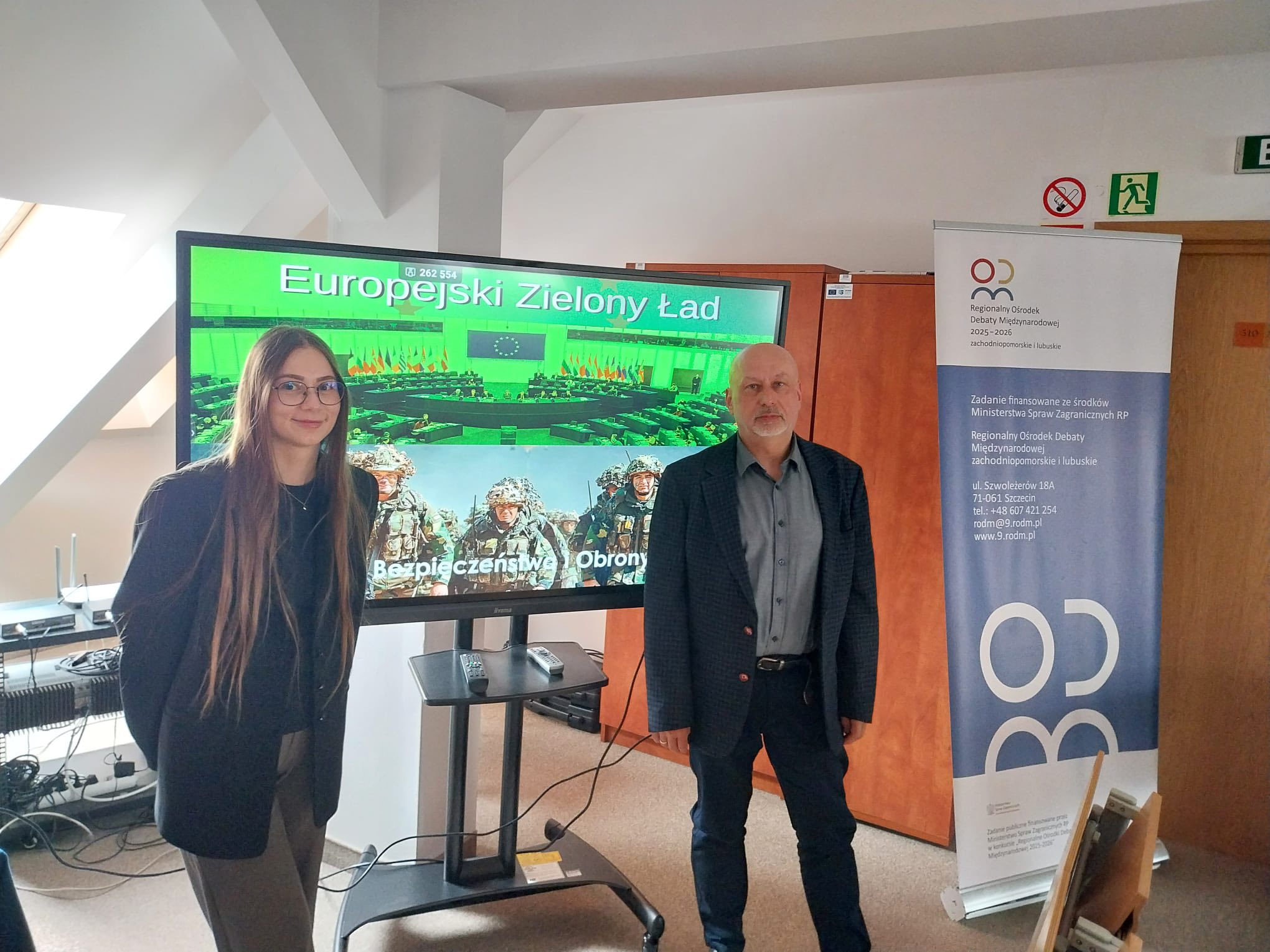 po prawej stoją: dr Arkadiusz Sójka, a po lewej mgr Oliwia Radkiewicz stoją podczas spotkania "O Unii przy kawie" z tyłu na telewizorze wyświetla się slajd dot. Europejskiego Ładu, a obok doktora Arkadiusza stoi roll-up Regionalnego Ośrodka Debaty Międzynarodowej