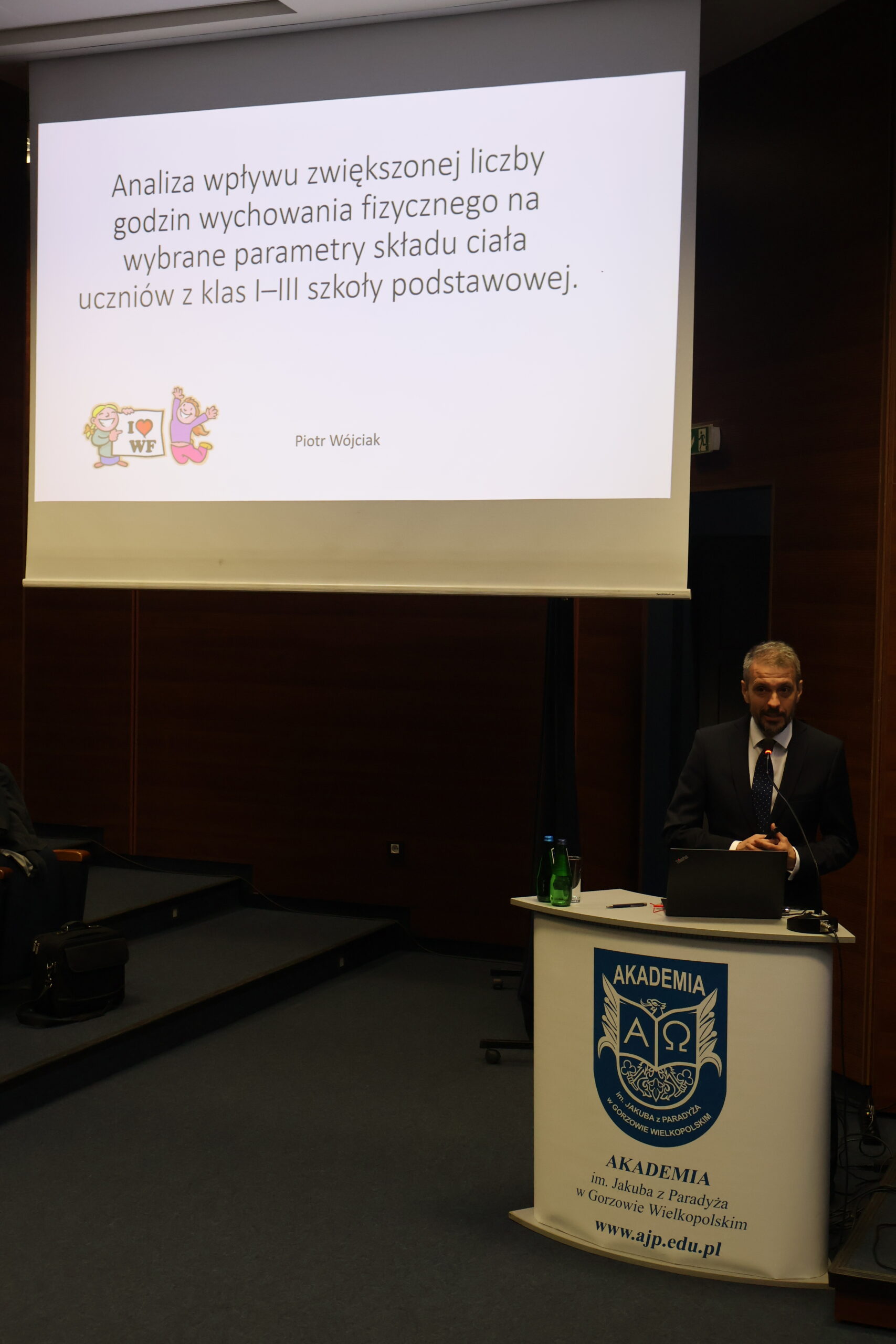 dr Piotr Wójciak stoi przy mównicy, w tle slajd z prezentacji