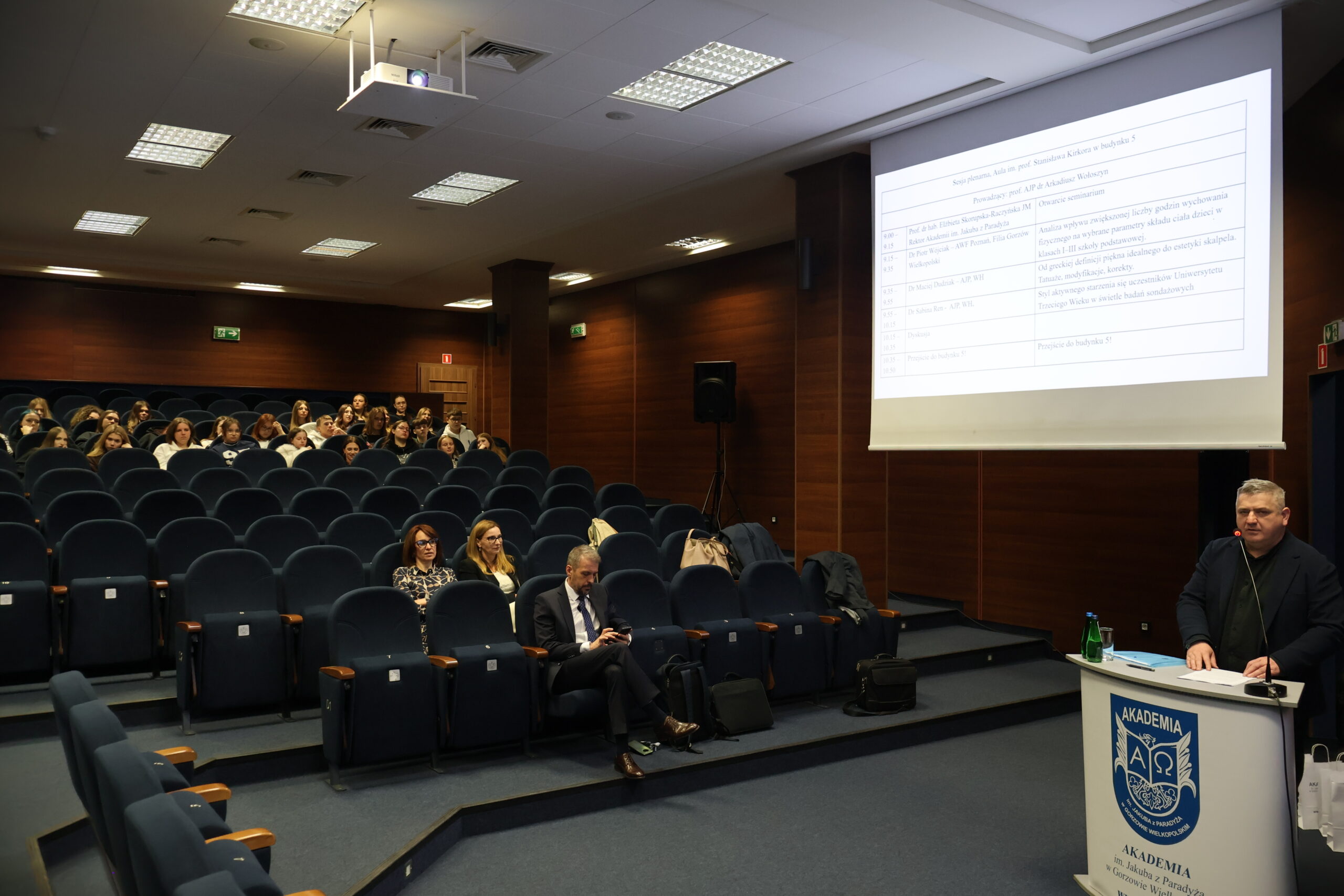 dr Maciej Dudziak stoi przy mównicy, w tle slajd z prezentacji