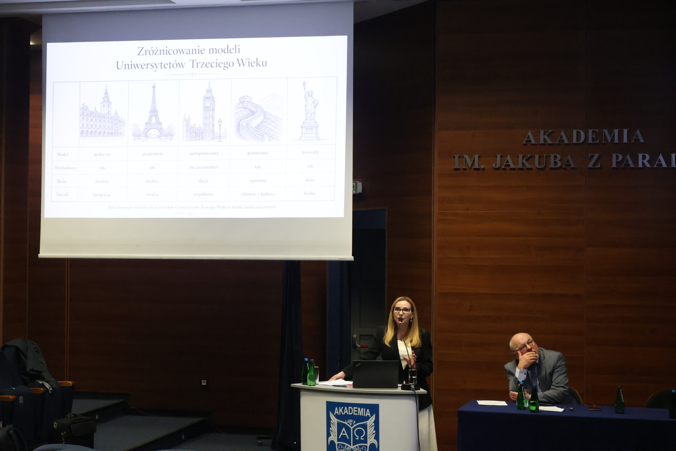 dr Sabina Ren stoi przy mównicy, obok siedzi prof. AJP dr Arkadiusz Wołoszyn, w tle slajd z prezentacji