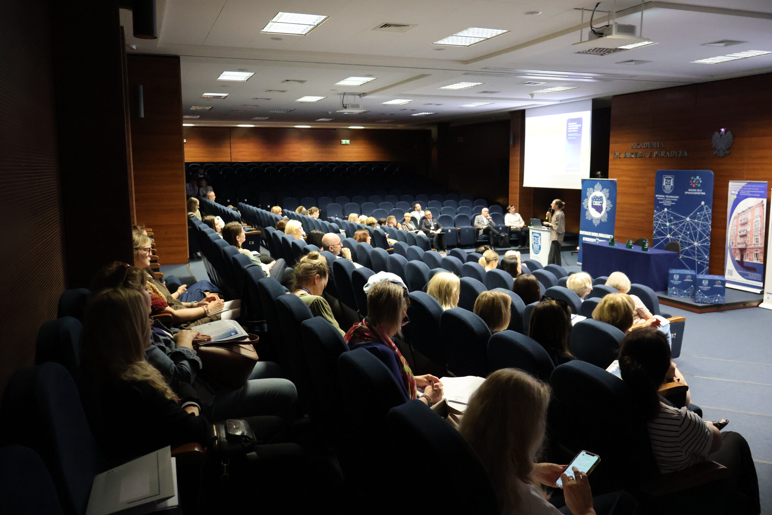 Uczestnicy konferencji siedzą, przed nimi prelegent wygłasza referat
