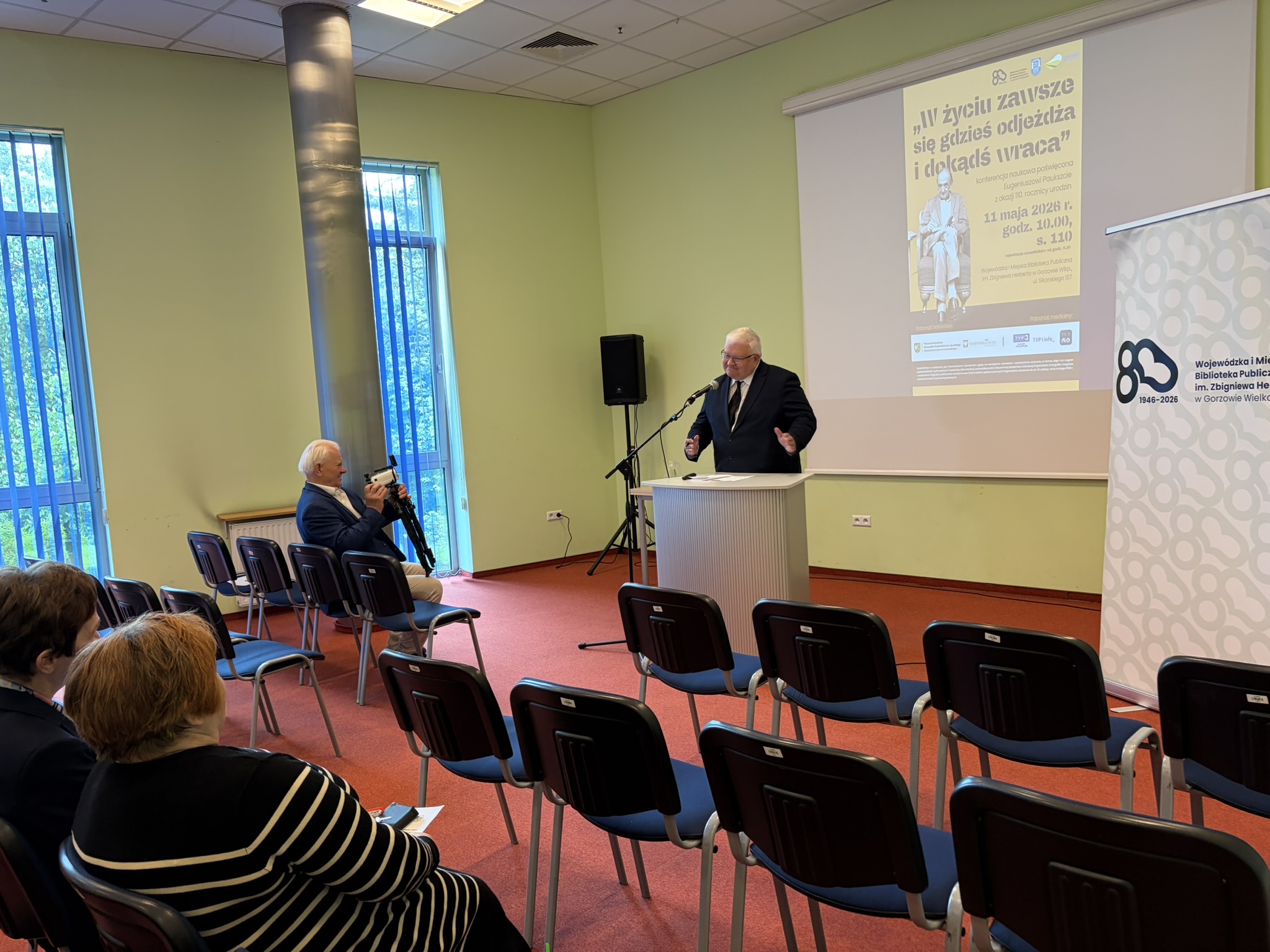 dr Sławomir Szenwaldprzemawia podczas Ogólnopolskiej Konferencji Naukowej z okazji 110. rocznicy urodzin Eugeniusza Paukszty. w tle siedzą zgromadzeni goście. Z tyłu wyświetla się slajd z plakatem odnośnie wydarzenia.