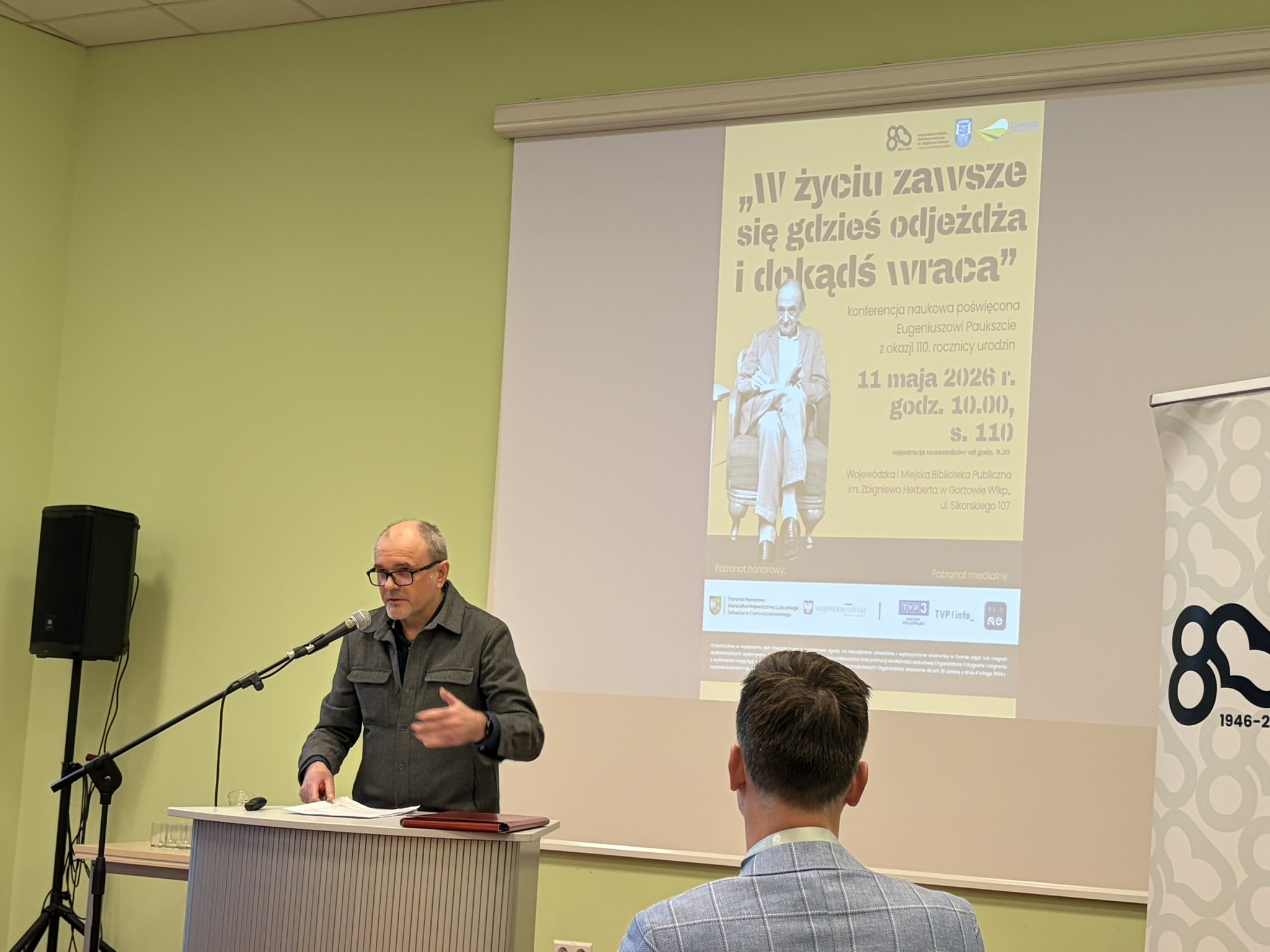 Prelegent podczas wygłaszania referatu podczas konferencji. Za nim w tle wyświetla się plakat dot. wydarzenia.