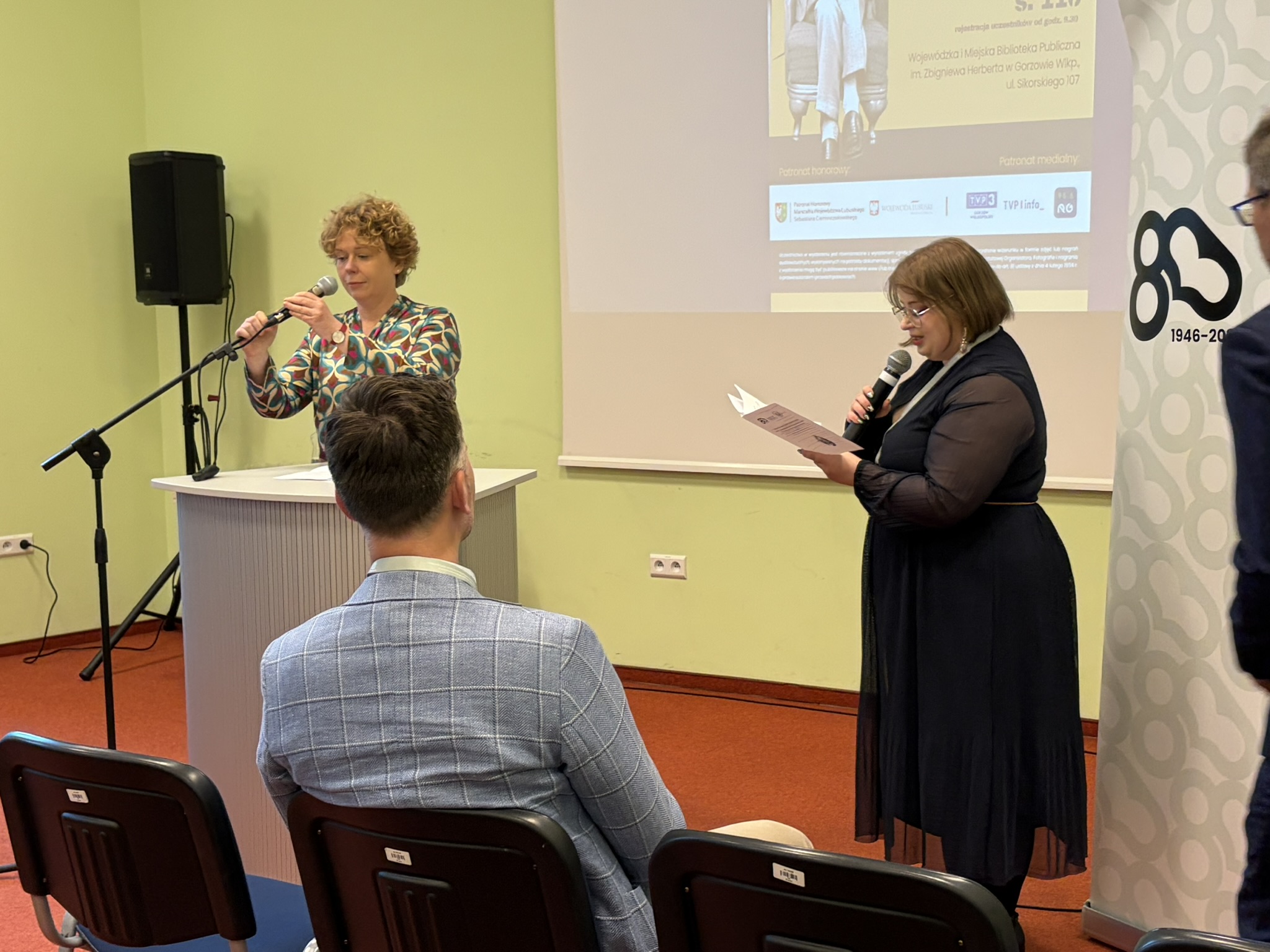 Koordynatorka spotkania zapowiada prelegentów, obok niej prelegentka przygotowuje sobie mikrofon do swojego wystąpienia.
