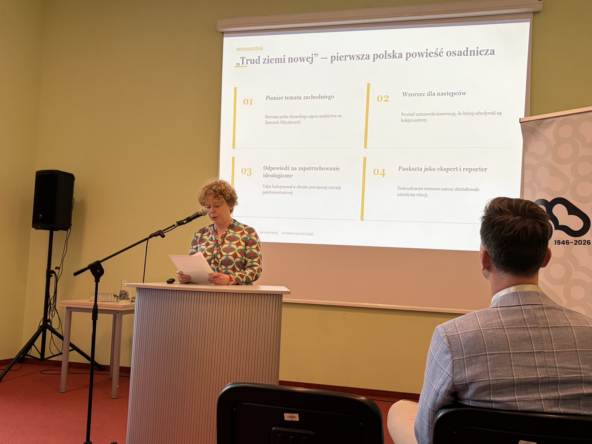 Prelegentka przy mównicy przemawia podczas Ogólnopolskiej Konferencji Naukowej z okazji 110. rocznicy urodzin Eugeniusza Paukszty. Za nią slajd "Trud ziemi nowej- pierwsza polska powieść osadnicza"