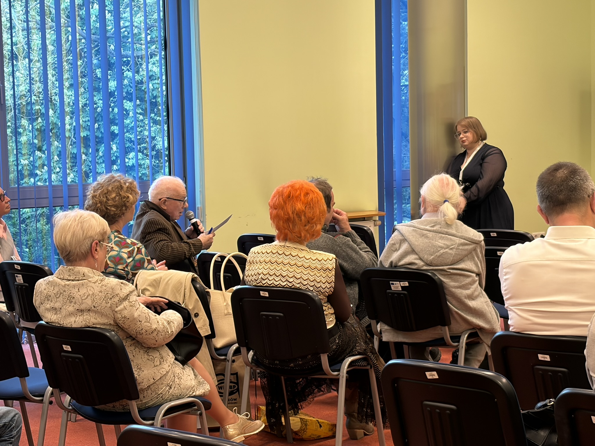 Mężczyzna z mikrofonem zabiera głos podczas dyskusji podczas Ogólnopolskiej Konferencji Naukowej z okazji 110. rocznicy urodzin Eugeniusza Paukszty. w tle siedzą zgromadzeni goście. Przy filarze stoi koordynatorka konferencji.